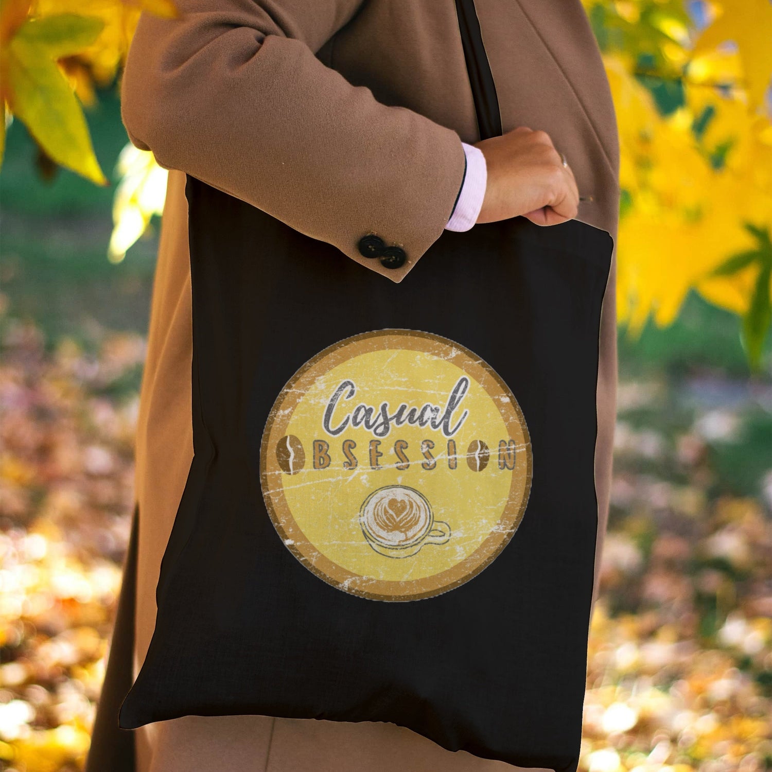 Autumn photo of a person in a leafy surrounding with a black canvas tote bag hanging from their shoulders with a Casual Obsession graphic image imprinted on one side.